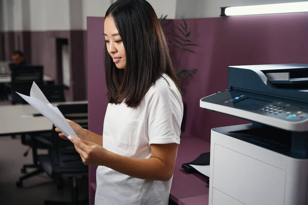 Side view portrait of stylish beautiful business woman with contract in hands is reading it while standing in the modern office