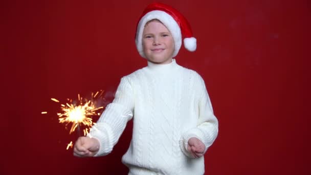 Menino feliz em Papai Noel cap segurando luz bengala no fundo vermelho — Vídeo de Stock