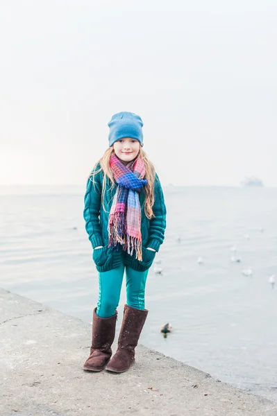 Portrait of a beautiful little girl on a cold weather next to lake, wearing, green jacket and pants, hat and scarf — Stock Photo, Image