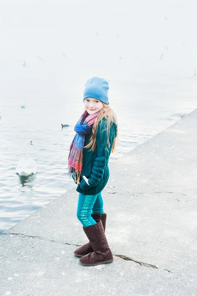 Portret van een mooi klein meisje op een koud weer naast lake, dragen, groene jas en broek, hoed en sjaal — Stockfoto