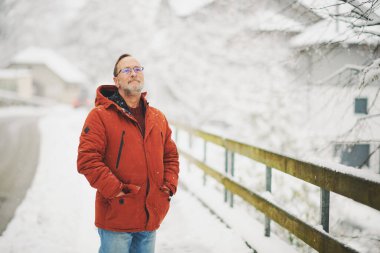 Orta yaşlı bir adamın açık hava portresi, 55-60 yaşlarında, soğuk bir günün tadını çıkarıyor, kırmızı turuncu kışlık ceket giyiyor.