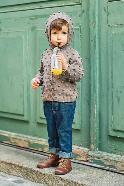 穿着棕色针织夹克 用吸管喝瓶子里的橙汁的可爱小女孩的户外肖像 — 图库照片