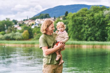 Genç ve mutlu bir babanın portresi. Küçük tatlı bir kızla gölün ya da nehrin kenarında oynuyor.