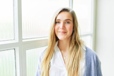 Büyük pencerenin yanında poz veren, doğrudan kameraya bakan 20 yaşında güzel bir kızın portresi.