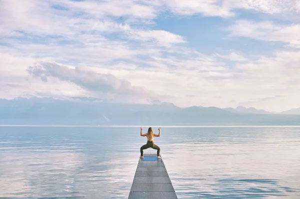 Outdoor portrait of young beautiful woman practicing yoga on dock by the lake, cactus pose, back view