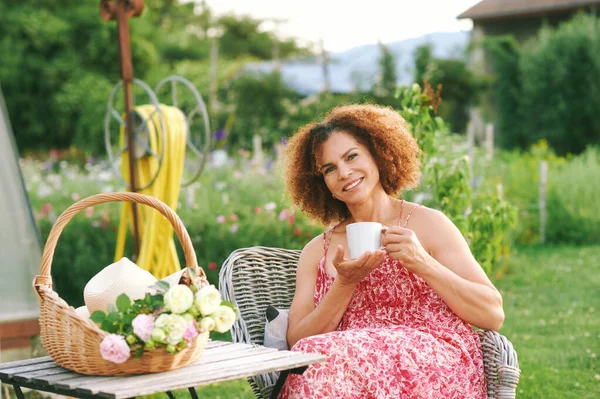 夏の庭で休んで美しい成熟した女性の屋外の肖像画 居心地の良い椅子に座って 紅茶やコーヒーのカップを保持し テーブルの上に立って新鮮なカットのバラとバスケット ロイヤリティフリーのストック写真
