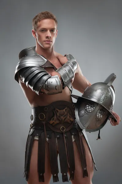 Gladiador em armadura posando com capacete sobre fundo cinza — Fotografia de Stock