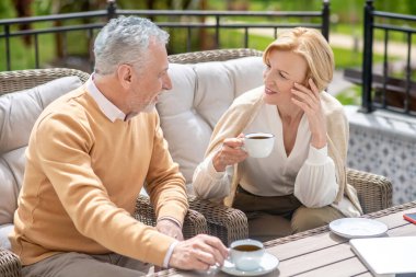 Koltuklarda oturmuş terasta konuşan iki orta yaşlı insan.