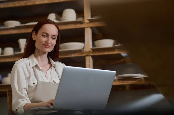 Uma jovem mulher sentada no laptop em uma oficina de cerâmica — Fotografia de Stock