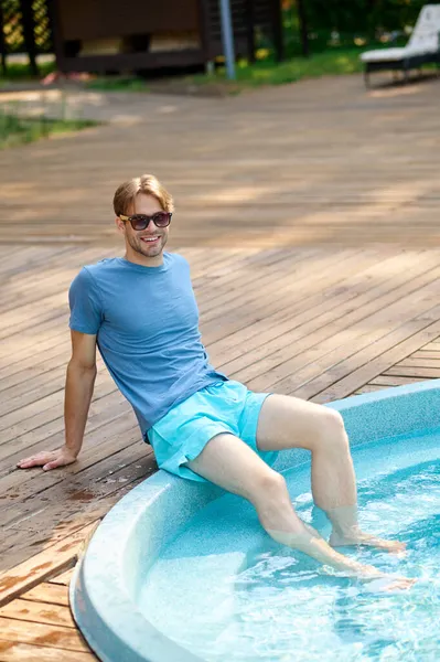 Ένας νεαρός άνδρας με μπλε μπλουζάκι και γυαλιά ηλίου διασκεδάζει στην πισίνα. — Φωτογραφία Αρχείου