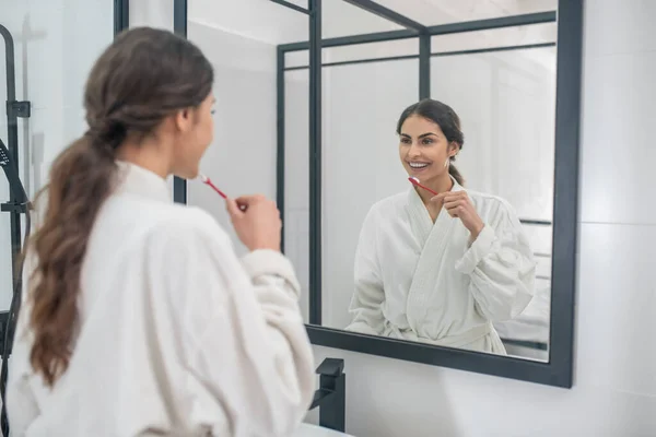Eine junge hübsche Frau im weißen Bademantel putzt sich die Zähne — Stockfoto