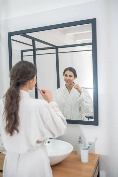 Eine junge hübsche Frau im weißen Bademantel putzt sich die Zähne — Stockfoto