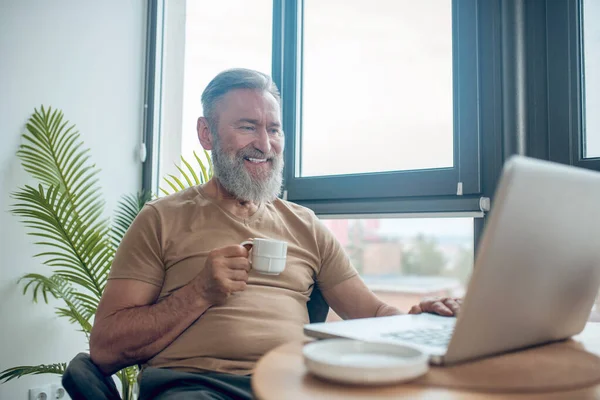 Um homem barbudo sentado no laptop wtahcing algo online e sorrindo — Fotografia de Stock