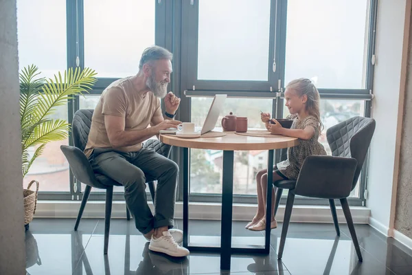 Un hombre y su hija sentados a la mesa, trabajando, su hija comiendo —  Fotos de Stock
