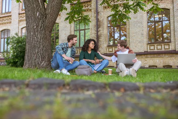 Junge Männer und ein Mädchen sitzen auf dem Rasen in der Nähe des College-Gebäudes — Stockfoto