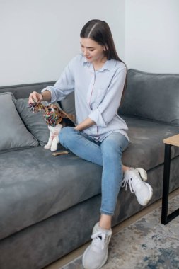 Genç, siyah saçlı bir kadın dizüstü bilgisayarda çalışıyor ve bir köpek yavrusuyla oynuyor.