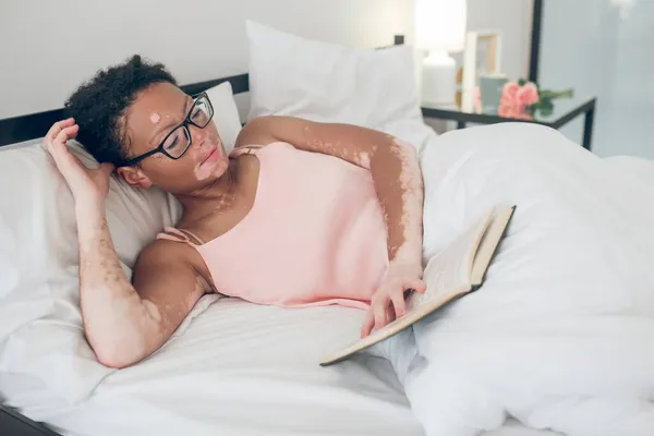 Μια νεαρή γυναίκα με γυαλιά διαβάζοντας ένα βιβλίο ενώ ξαπλώνει στο κρεβάτι — Φωτογραφία Αρχείου