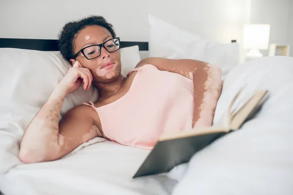 Seorang wanita muda berkacamata membaca buku saat berbaring di tempat tidur — Stok Foto