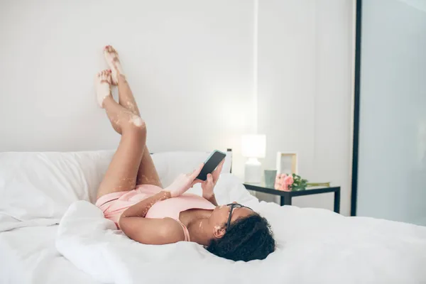 Женщина в нижнем белье лежит в постели с поднятыми ногами и смотрит что-то на смартфоне — стоковое фото