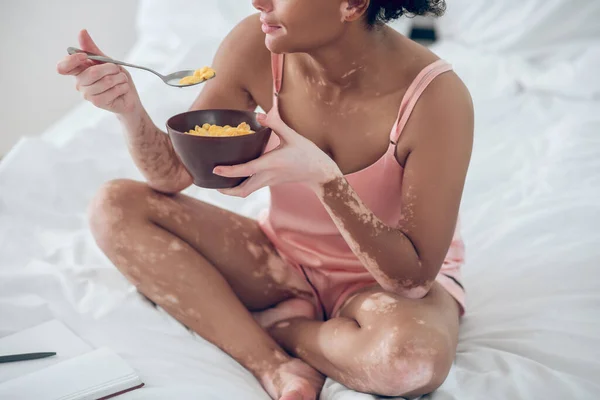 Een donkere vrouw die ontbijtgranen eet terwijl ze op bed zit — Stockfoto