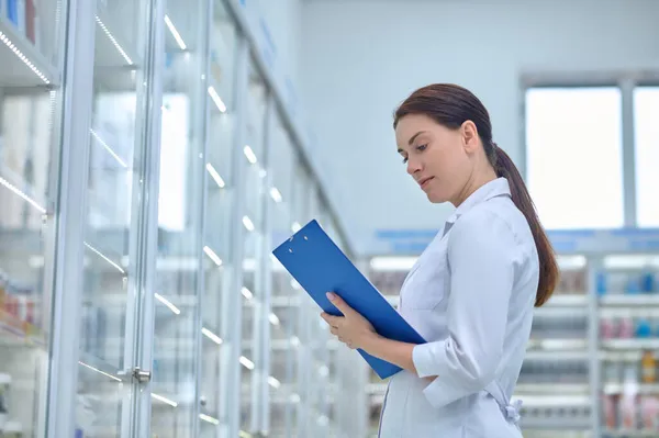 Mujer tomando notas cerca de los estantes de medicamentos en la farmacia —  Fotos de Stock