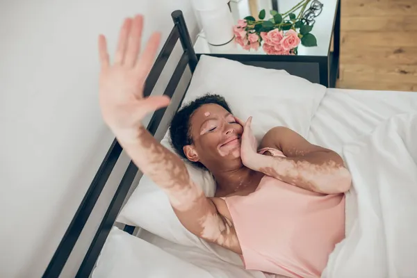 Uma jovem mulher de pele escura deitada na cama, estendendo a mão e sorrindo — Fotografia de Stock