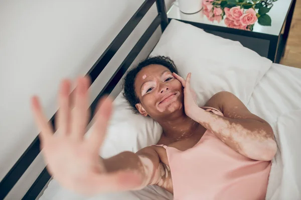 Egy fiatal, sötét bőrű nő fekszik az ágyban, kinyújtott kézzel és mosolyogva. — Stock Fotó
