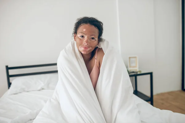 Um sorrindo bonito jovem mulher sentada na cama coberta com cobertor — Fotografia de Stock