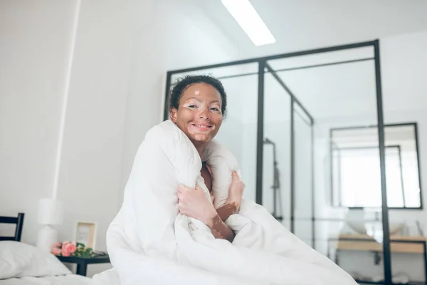 Улыбающаяся симпатичная молодая женщина, сидящая на кровати, покрытой одеялом — стоковое фото