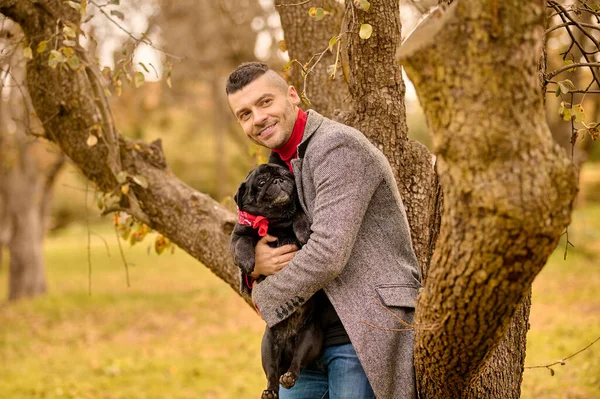 Ένας άντρας στέκεται κοντά στο δέντρο με ένα σκύλο στα χέρια. — Φωτογραφία Αρχείου