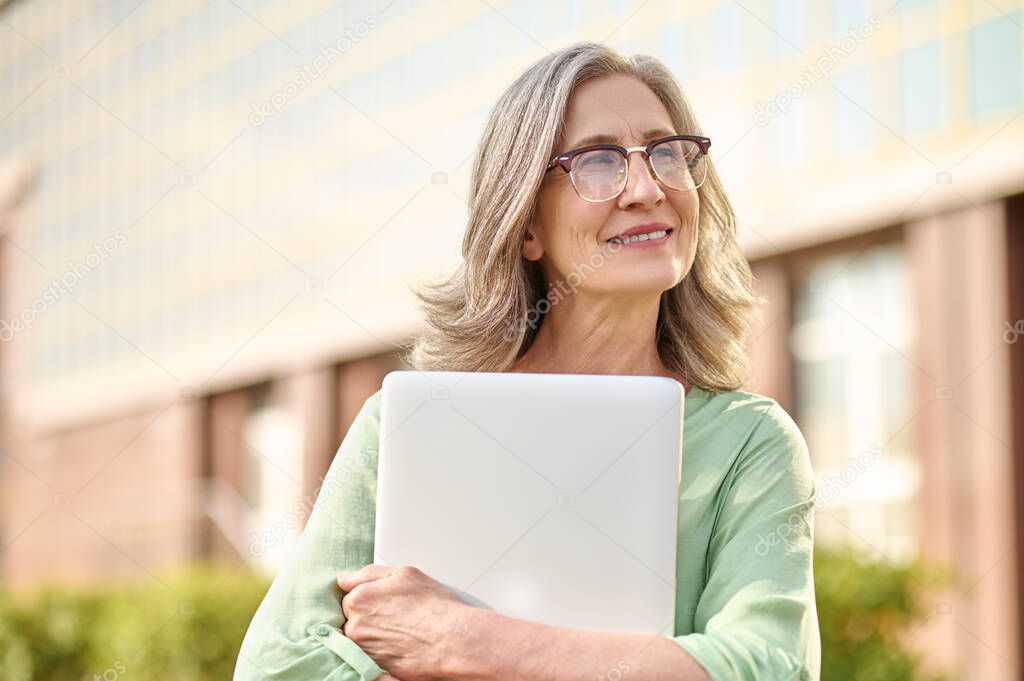 Thoughtful woman in glasses hugging laptop on street