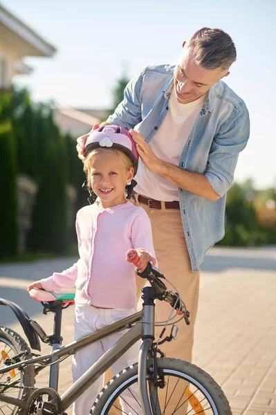 A man putting protective helmet on his daughters head before riding a bike — Stock Photo, Image