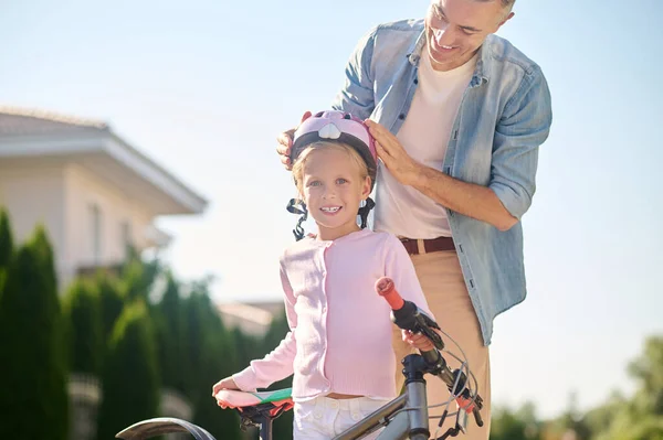 Un hombre que pone casco protector en la cabeza de sus hijas antes de montar en bicicleta — Foto de Stock