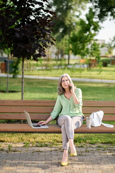 Женщина с ноутбуком на скамейке говорит на смартфоне — стоковое фото