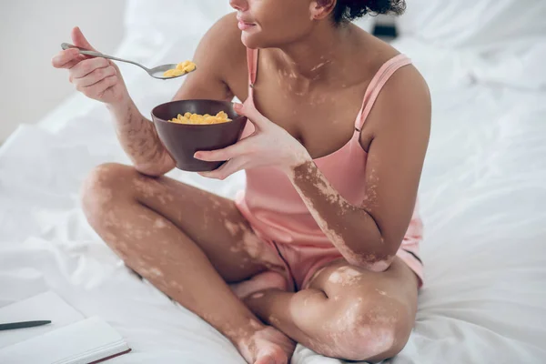 En mörkhyad kvinna äter flingor medan hon sitter på sängen — Stockfoto