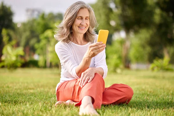 Женщина сидит на траве и смотрит в смартфон — стоковое фото