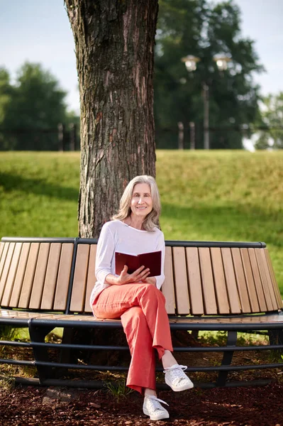 Kvinna med bok sittande på bänk under träd — Stockfoto