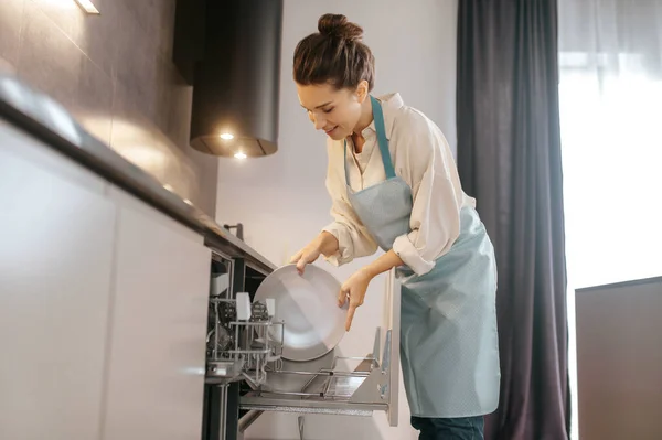 Женщина, стоящая возле посудомоечной машины и вынимающая тарелки — стоковое фото