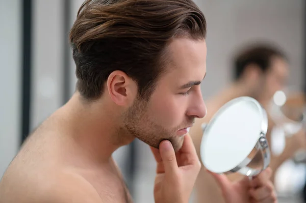 Młody człowiek badający swoją twarz patrząc w okrągłe lustro — Zdjęcie stockowe