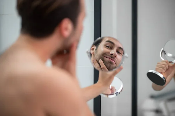 Jongeman onderzoekt zijn gezicht terwijl hij in de ronde spiegel kijkt — Stockfoto