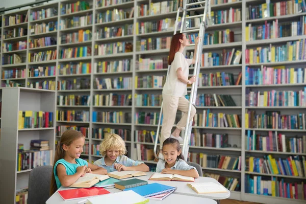 Children reading at table and woman on ladder near bookshelf — 图库照片