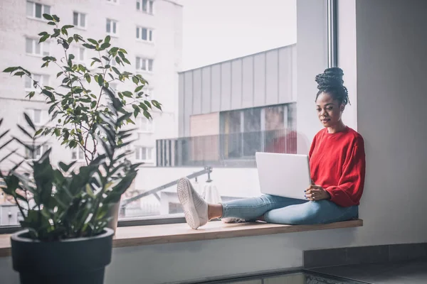 Fille à la peau foncée assise sur le rebord de la fenêtre du bureau avec ordinateur portable. — Photo