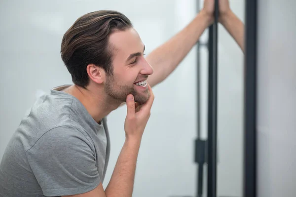 Een jongeman in grijs T-shirt kijkt naar zijn spiegelbeeld in de spiegel — Stockfoto