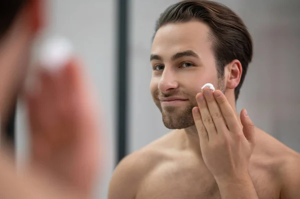 一个半裸的男人站在镜子旁边，把剃须泡沫涂在他的脸上 — 图库照片