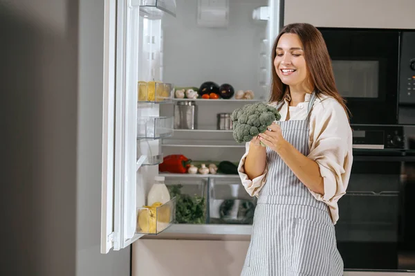 Jonge huisvrouw staat bij de koelkast in de keuken — Stockfoto