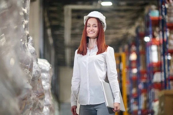 Woman in helmet with laptop standing in aisle of warehouse — Zdjęcie stockowe