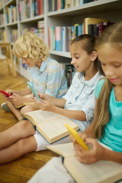 Boy and two girls looking at smartphones in library — Stock Fotó