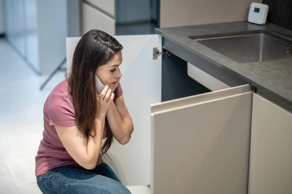 Mujer confundida con teléfono inteligente cerca de la oreja mirando debajo del fregadero — Foto de Stock