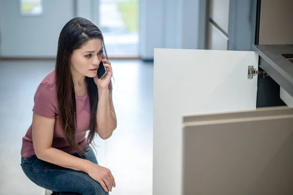 Woman crouching near kitchen sink with smartphone near ear