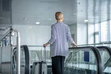Elegant female tourist with the trolley suitcase riding the escalator